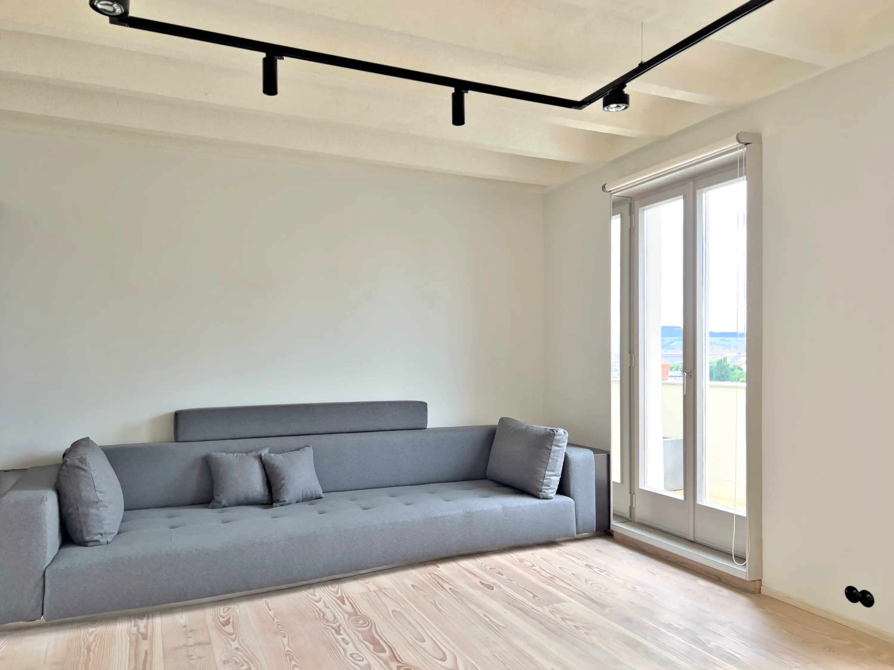 Großzügiger, lichtdurchfluteter Wohnraum mit schlichtem grauen Sofa und bodentiefen Fenstern.