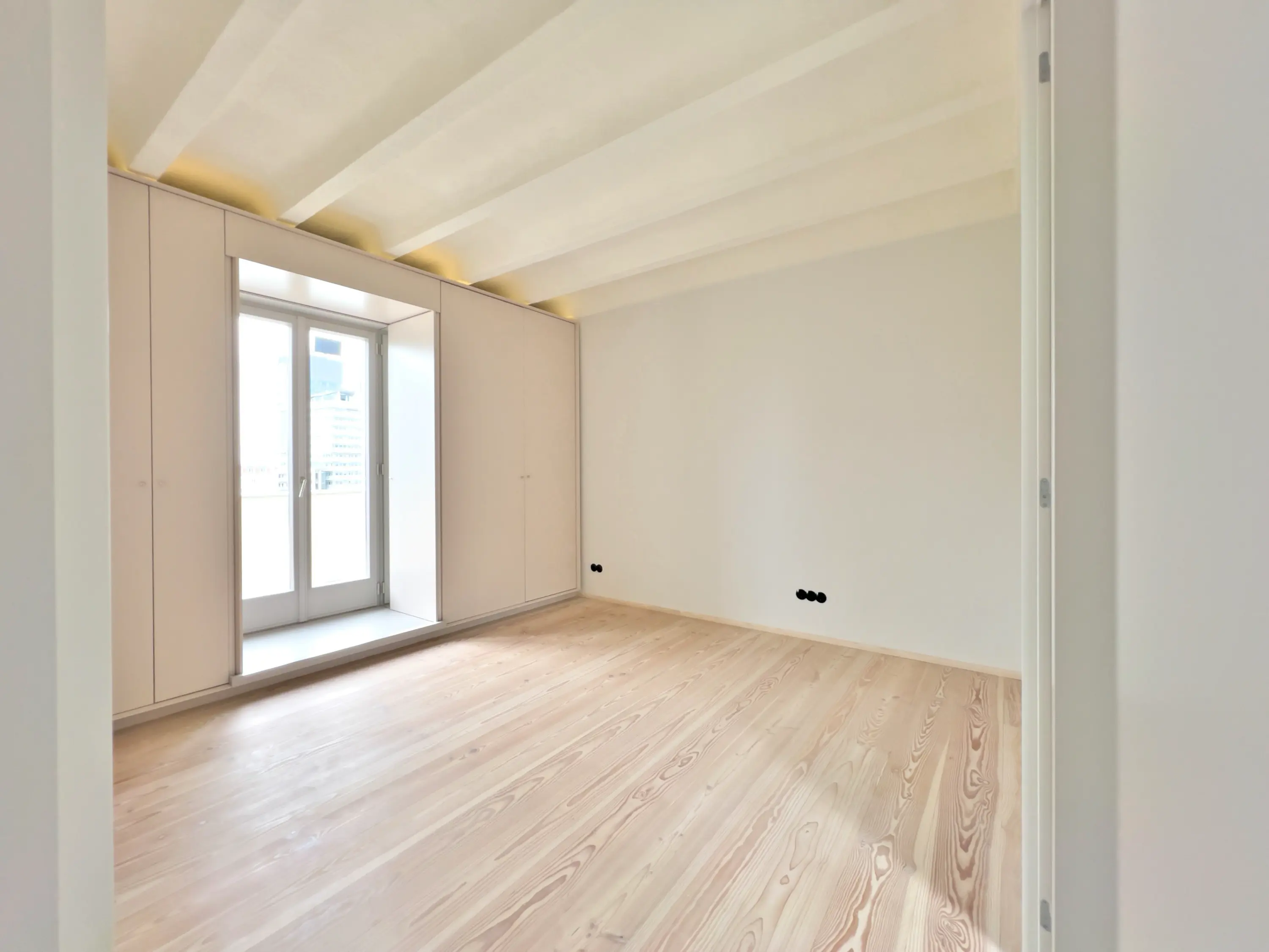 Weitläufiger Raum mit industrieller Deckenstruktur, großen Fenstern und hellem Holzfußboden.